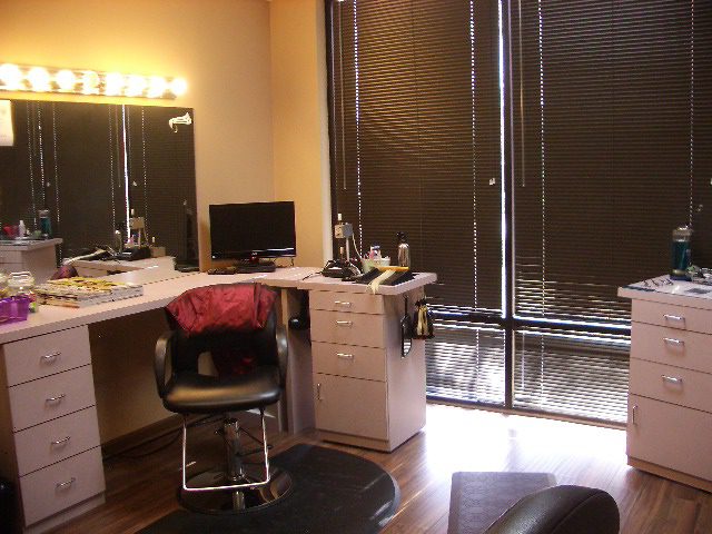 Hair salon room with a styling chair, vanity mirrors, and a television. Countertops hold hair products and tools. Blinds cover the large windows, and the lighting is warm.