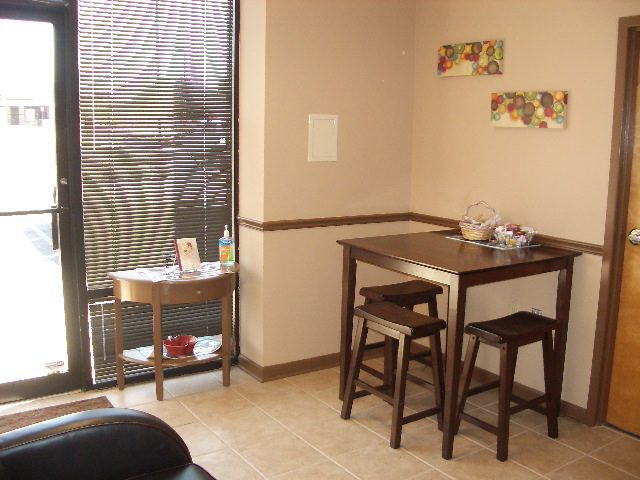 A small room with a high table and two stools, a side table by the window with blinds, and colorful wall art.