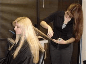 A woman is seated in a salon chair having her long, blonde hair trimmed by a hairstylist using scissors.